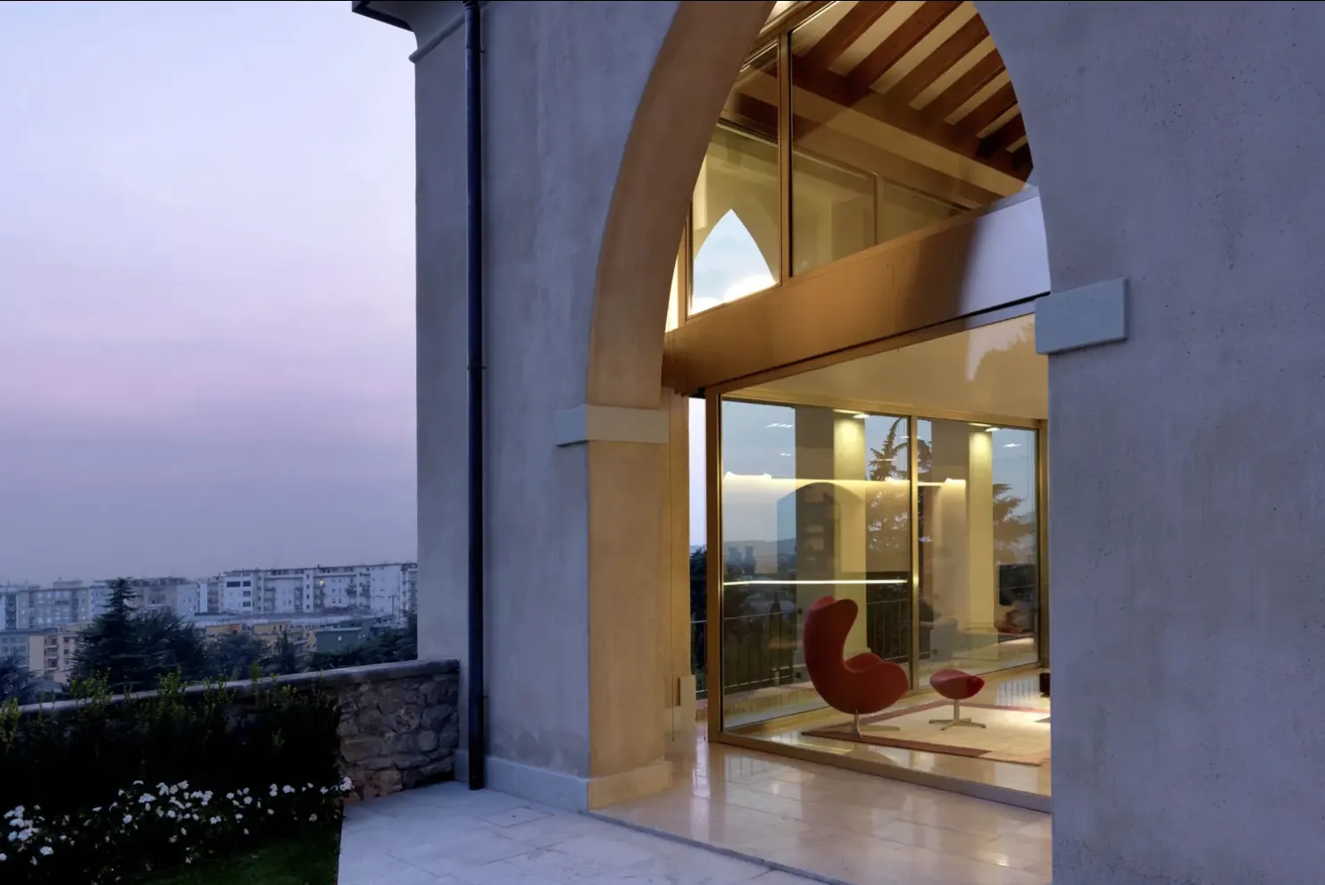 Grande arco in pietra chiuso da una vetrata: all'interno travi in legno e una poltrona rossa, all'esterno la città al tramonto