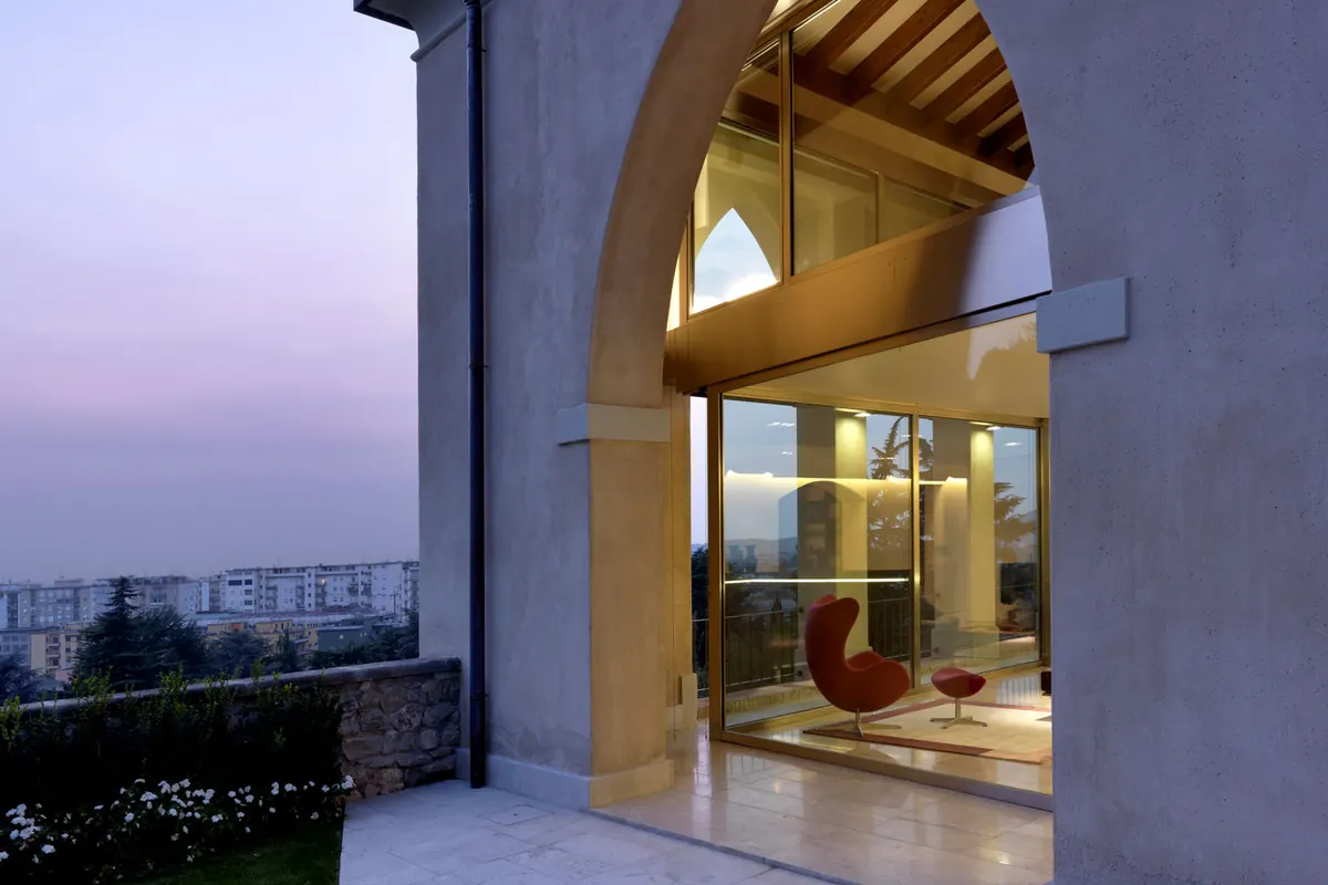 Arco in pietra chiuso da una vetrata al tramonto, con la poltrona rossa all'interno e la città sullo sfondo