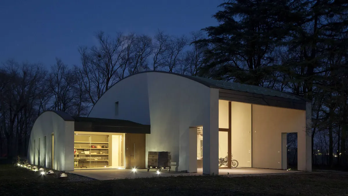 Villa in campagna di sera: il volume bianco con la copertura curva, la grande vetrata illuminata e gli alberi alle spalle