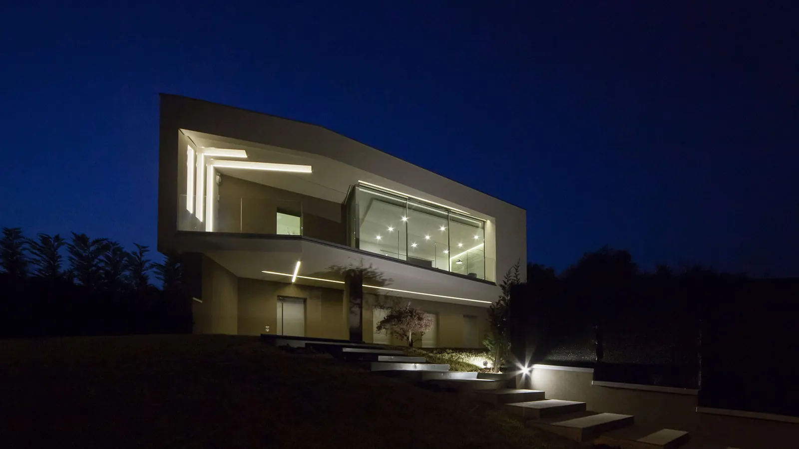 Villa sul Lago di Garda di notte: volume bianco a sbalzo con la loggia vetrata illuminata e la scala in pietra nel prato
