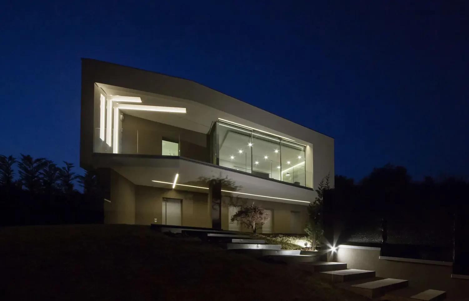 Villa contemporanea di notte: volumi bianchi a sbalzo, vetrate illuminate e gradini di luce nel prato