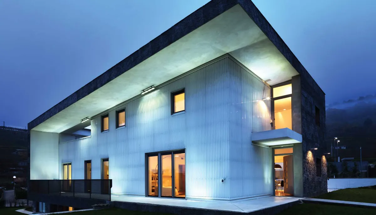 Villa a due piani al crepuscolo: il volume chiaro con la copertura a sbalzo illuminata dal basso e le finestre accese