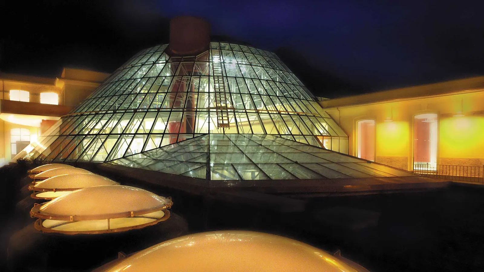 La cupola tronco-conica in vetro illuminata di notte, con i lucernari a calotta in primo piano e i corpi dell'ex cotonificio intorno