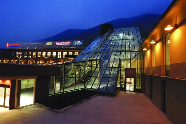 La galleria vetrata a gradoni illuminata di sera, tra il corpo storico con le insegne e la facciata ocra, con le montagne sullo sfondo