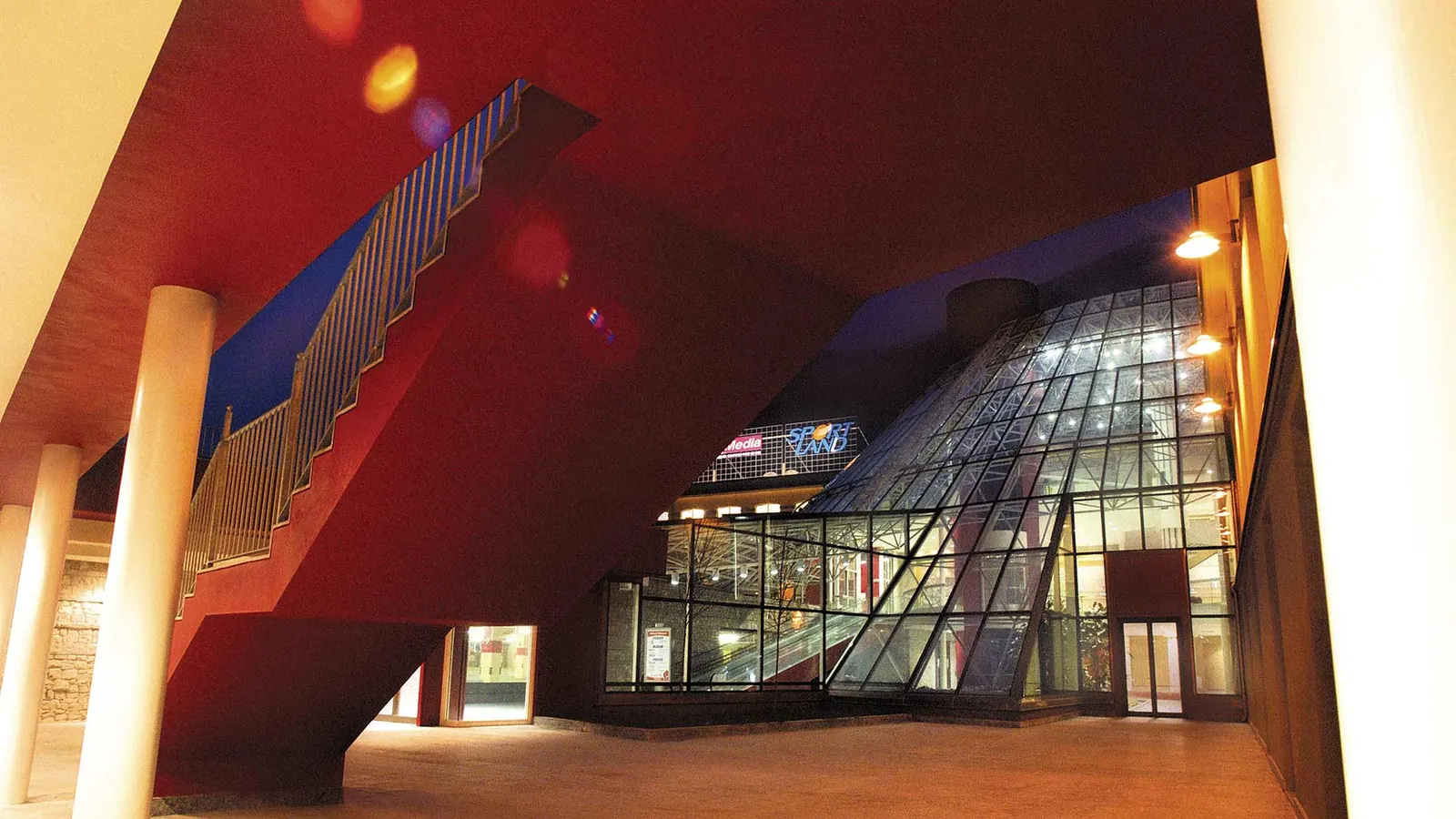 Portico rosso con scala esterna e colonne chiare; sullo sfondo la galleria vetrata illuminata, di sera