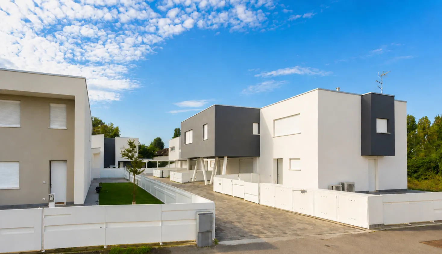 Residenze Theico: volumi bianchi e grigi con recinzioni chiare lungo una strada privata, sotto un cielo con nuvole