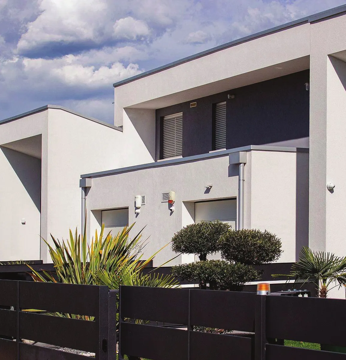 Theico: residenze a volumi bianchi squadrati con recinzioni scure e piante, sotto un cielo con nuvole