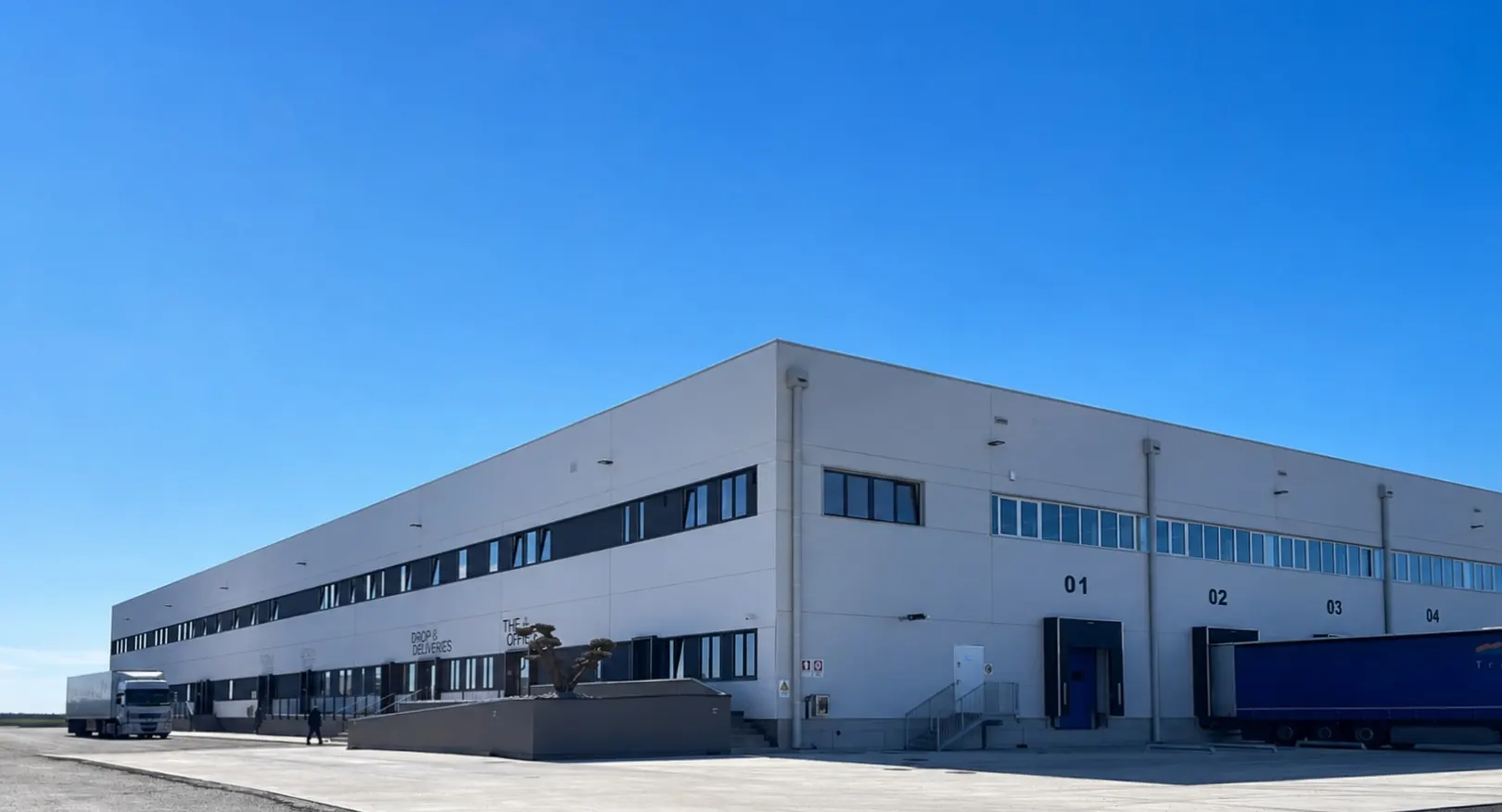 Fronte del magazzino Transbozen con le baie di carico, un semirimorchio in sosta e il cielo terso