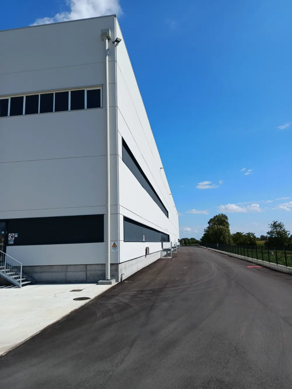 Fronte laterale del magazzino: parete bianca con le finestre a nastro, la strada di servizio e il cielo blu