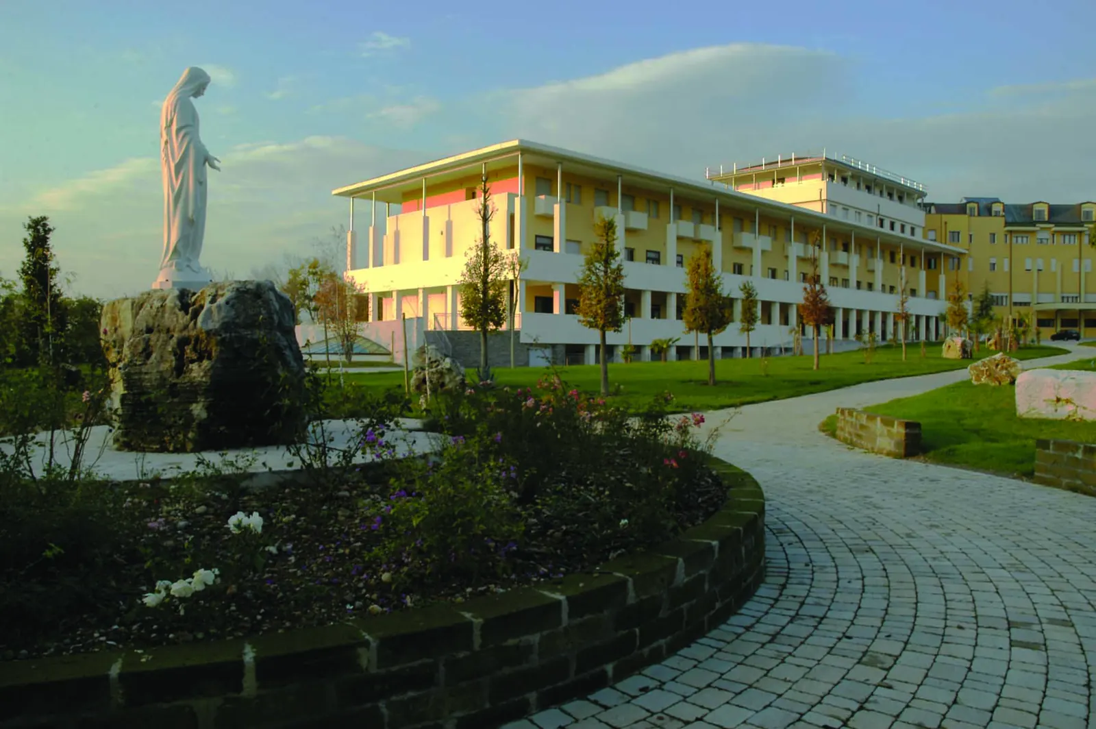 Comunità missionaria di Villaregia: la statua della Madonna nel giardino e, dietro, il nuovo edificio con il lungo portico, al tramonto